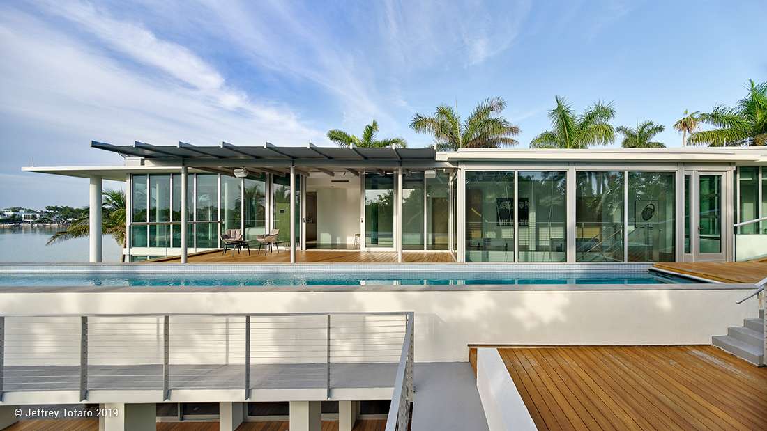 Modern waterfront home featuring flat roof design, floor-to-ceiling glass walls, sliding glass doors, and outdoor deck with infinity-edge pool.