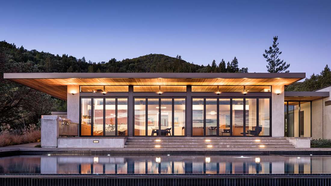 Luxury modern home with flat roof design, floor-to-ceiling glass walls, illuminated interior, and outdoor infinity-edge pool reflecting warm lights against a backdrop of forested hills at dusk.