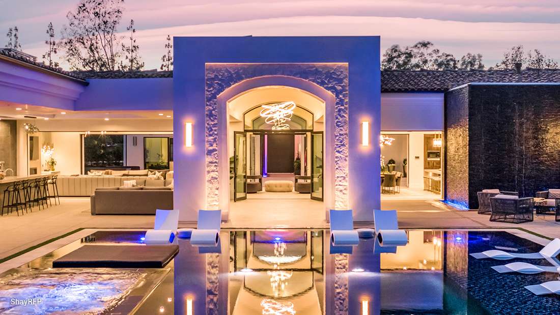 Luxury modern home with illuminated arched entryway, textured stone facade, and open-concept interior featuring lounge seating and bar area, overlooking a sleek infinity pool with submerged loungers at dusk.