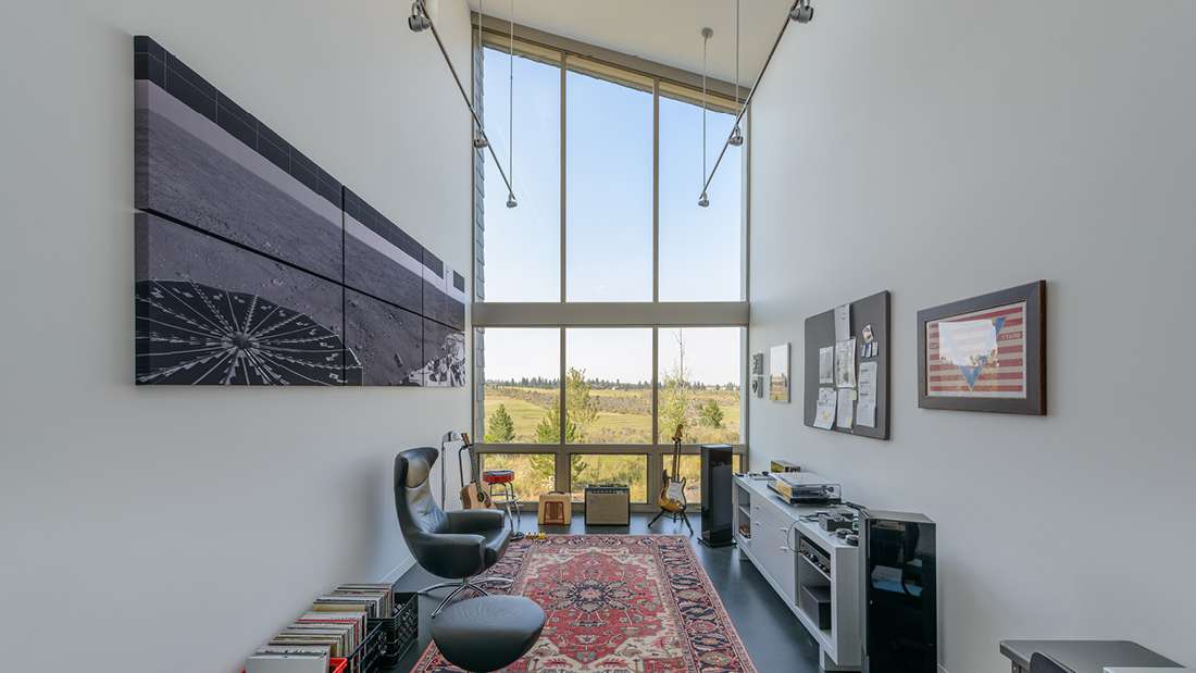 Modern music room with vaulted ceiling, floor-to-ceiling glass windows offering panoramic countryside views, black leather lounge chair, contemporary artwork, and red patterned area rug.