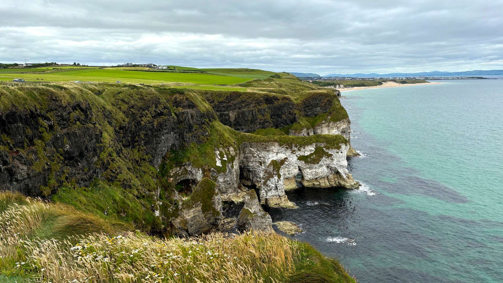 Steep grassy cliffs with white rock faces overlooking turquoise ocean under a cloudy sky along rugged coastline.