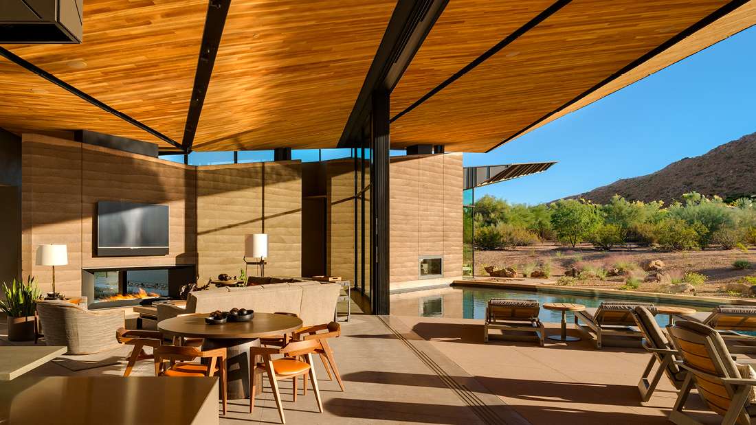 Luxury desert home with open-concept living area, wood-paneled ceiling, modern fireplace, and sliding glass walls opening to a poolside terrace with lounge chairs and mountain views.