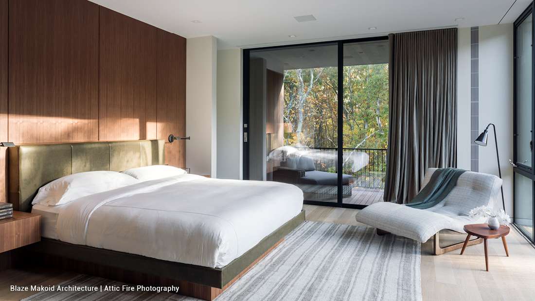 Modern bedroom with wood-paneled accent wall, upholstered headboard, minimalist decor, and sliding glass doors opening to a private balcony with forest views.