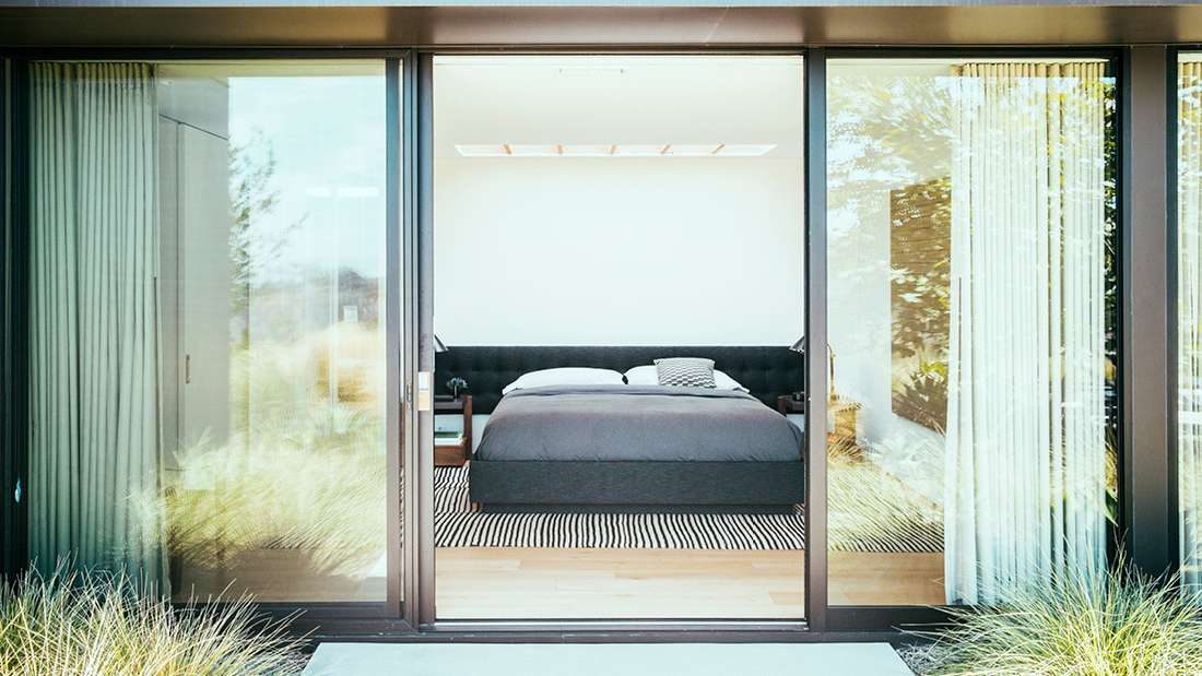 Minimalist bedroom with black upholstered bed, striped area rug, and large sliding glass doors opening to outdoor greenery.