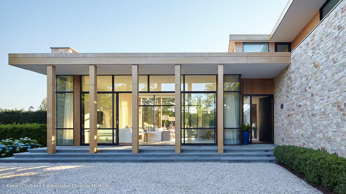 Modern luxury home exterior with stone accent wall, vertical wooden columns, and floor-to-ceiling glass windows framed by sleek black trim.