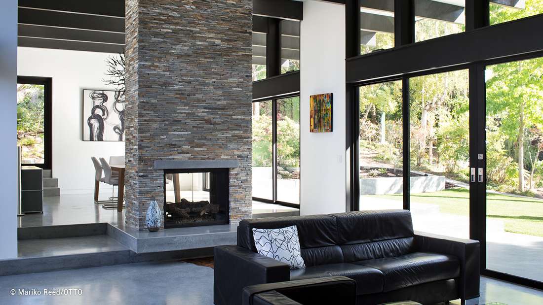 Modern living room with stacked stone fireplace, black leather sectional sofa, and floor-to-ceiling glass doors opening to a landscaped garden.
