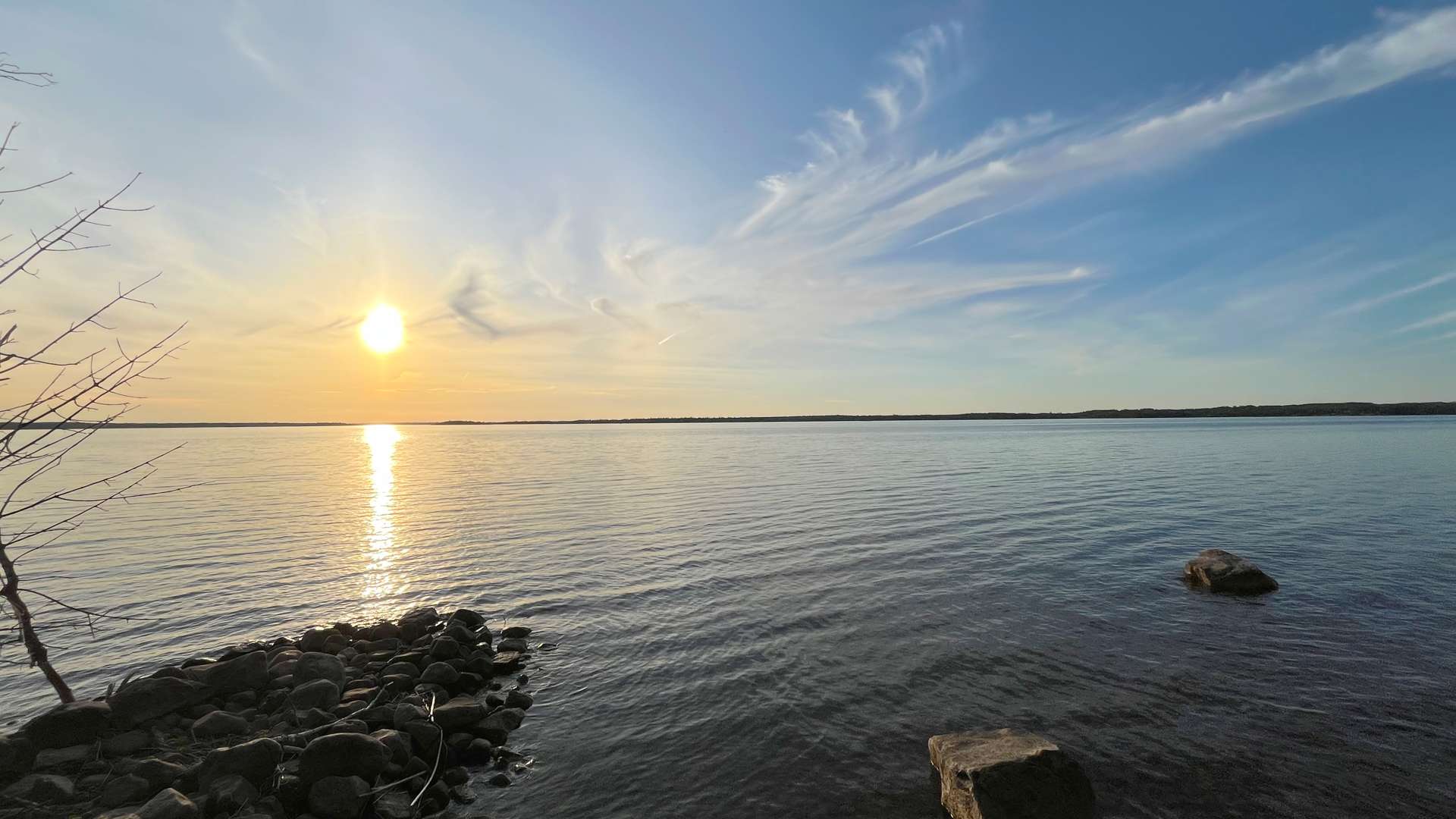 Calm lake at sunset with golden sunlight reflecting on the water, scattered clouds in a blue sky, and rocks along the shoreline.