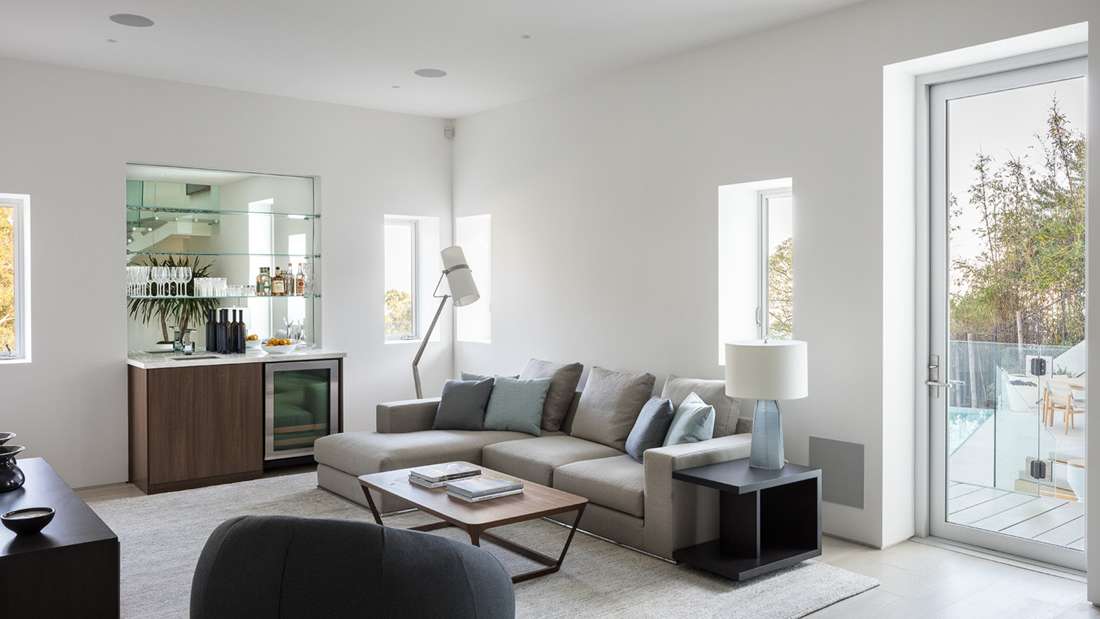 Bright modern living room with sectional sofa, mirrored bar cabinet, glass shelving, and floor-to-ceiling glass door opening to outdoor patio with pool.