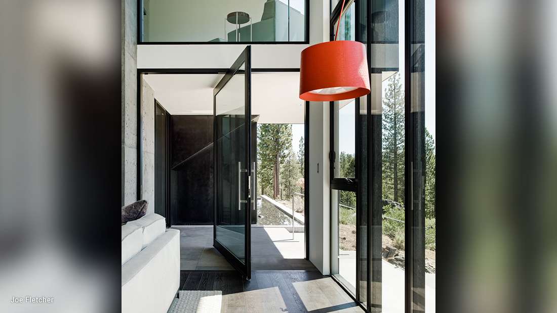 Modern entryway with pivot glass door framed by black metal, floor-to-ceiling windows offering forest views, minimalist concrete flooring, and bold red pendant light fixture.