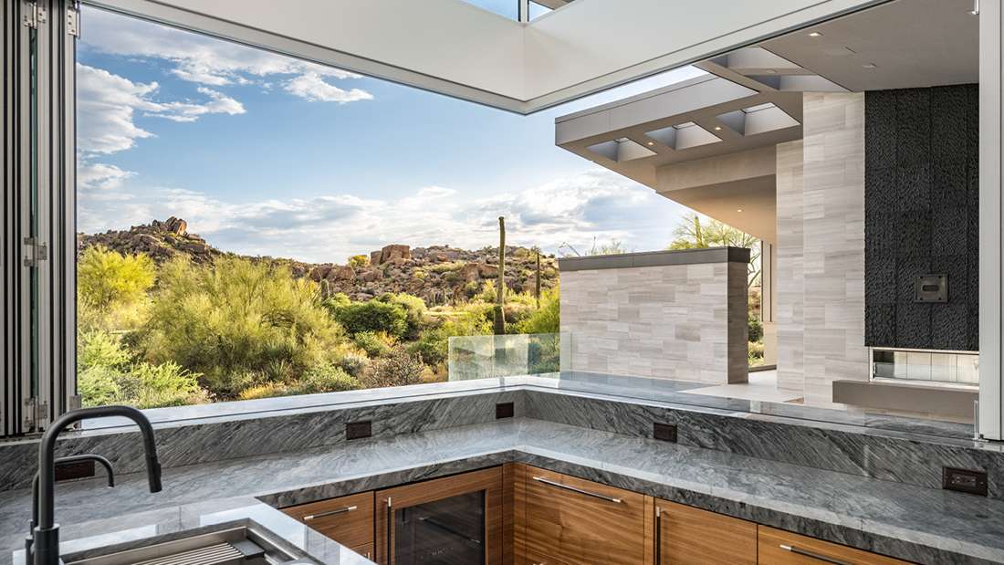Luxury modern kitchen with open corner design, gray marble countertops, light wood cabinetry, and folding glass windows offering panoramic desert views and natural light.