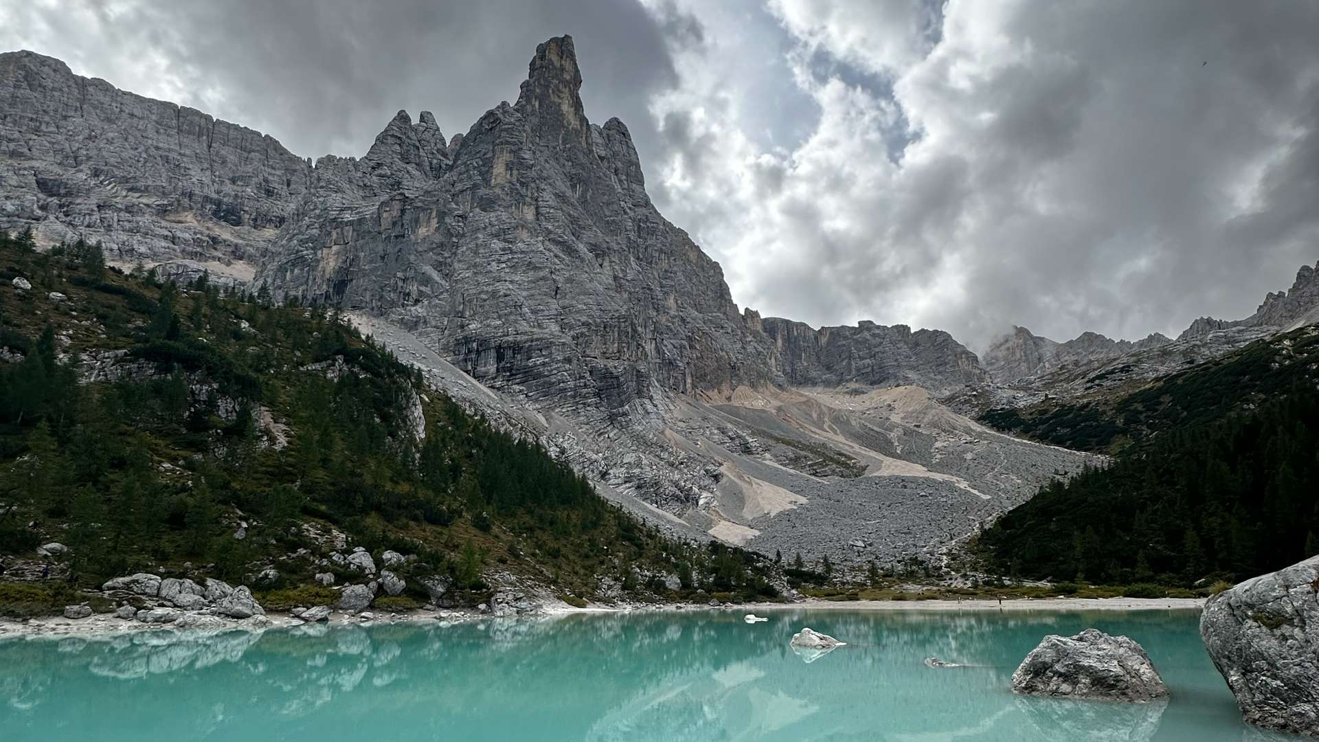 Turquoise alpine lake surrounded by rocky terrain and dense green forest, with towering jagged mountain peaks under dramatic cloudy skies in the Dolomites.