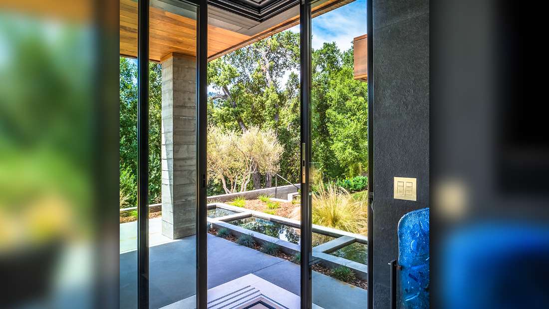 Modern sliding glass doors opening to a covered patio with concrete flooring, wooden ceiling, and landscaped garden featuring lush greenery and water feature.