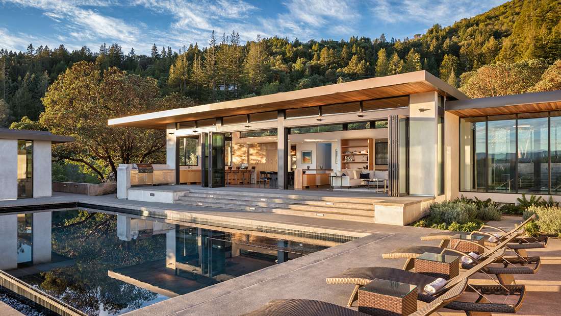 Luxury modern hillside home with flat roof design, floor-to-ceiling glass walls, open-concept living area, and outdoor terrace featuring infinity-edge pool and lounge chairs overlooking forested mountains.