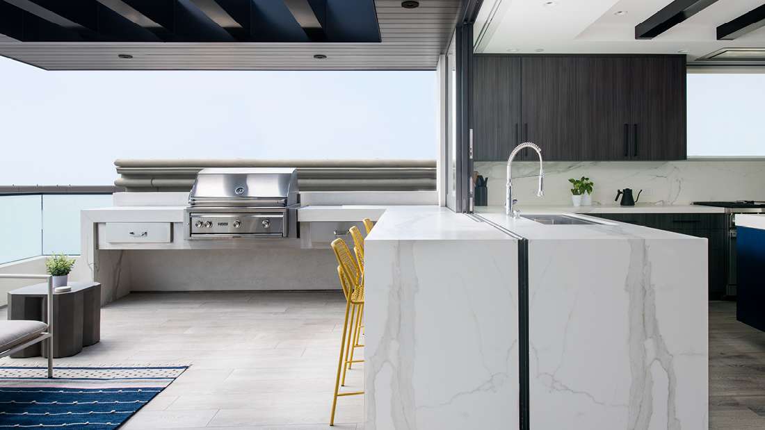 Modern outdoor kitchen with built-in stainless steel grill, marble waterfall island with integrated sink, yellow bar stools, and sleek cabinetry under a slatted pergola.