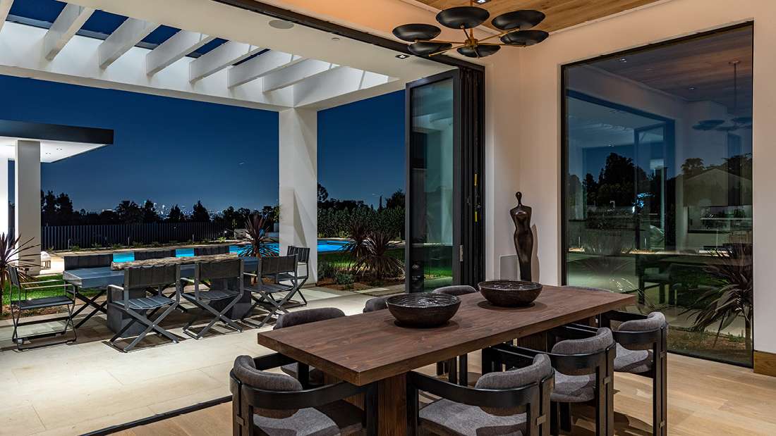 Luxury indoor dining area with dark wooden table and upholstered chairs, folding glass doors opening to outdoor patio with pergola, modern outdoor dining set, and illuminated swimming pool under night sky.