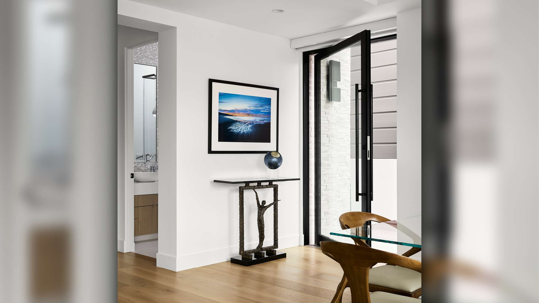Modern interior entryway with black-framed glass pivot door, white walls, light wood flooring, minimalist console table with sculptural base, framed ocean photograph, and glimpse of adjacent bathroom with mosaic tile wall.