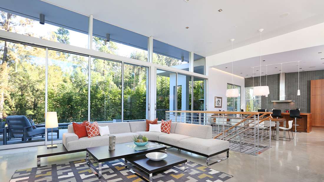 Open-concept modern living room with floor-to-ceiling glass walls, forest views, light gray sectional sofa, and sleek kitchen with pendant lighting.