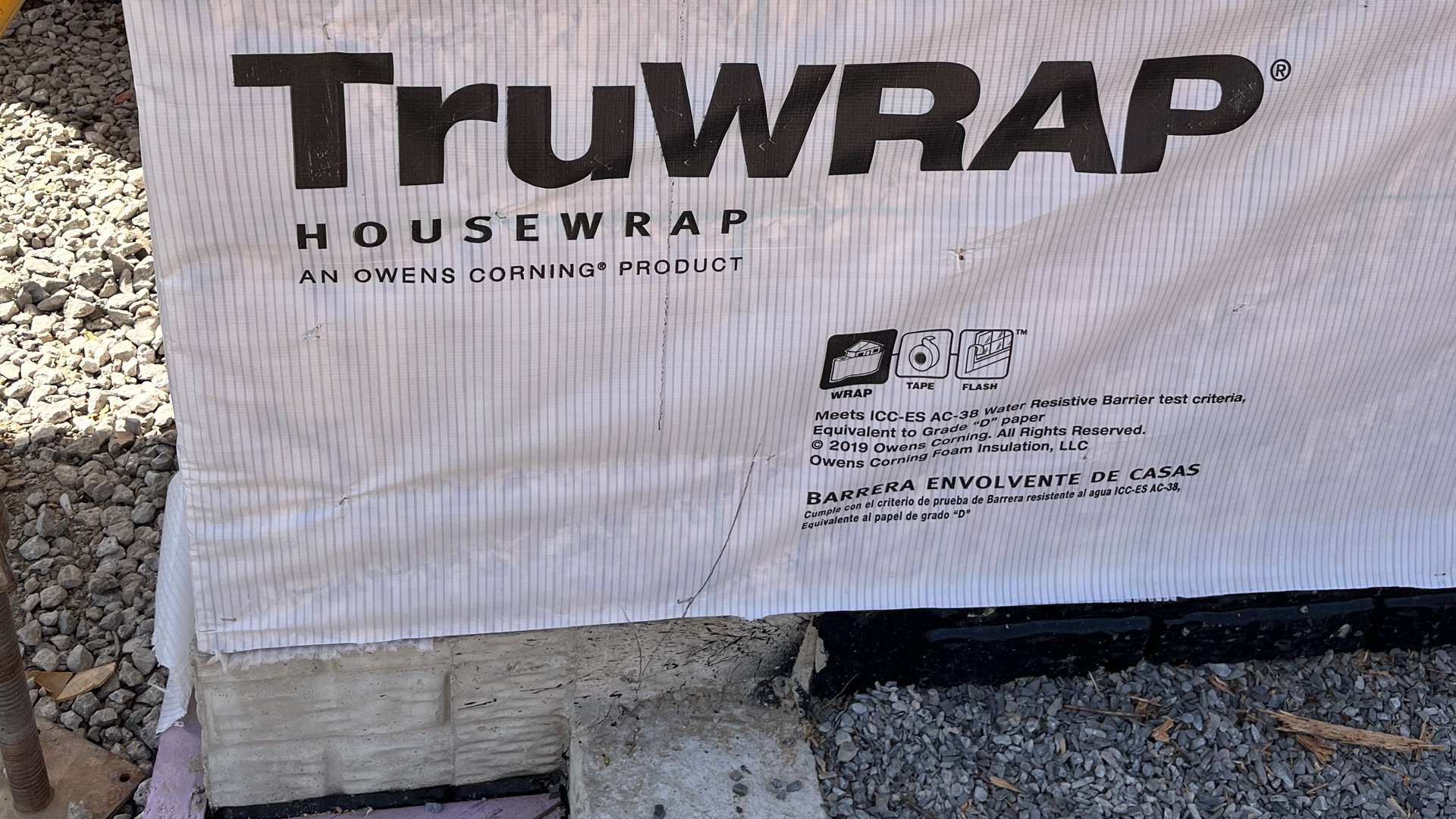 Close-up of Owens Corning TruWrap housewrap installed on a foundation wall at a construction site with gravel ground.