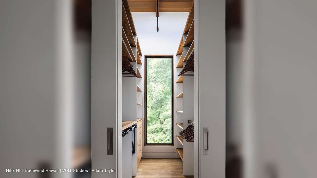 Modern walk-in closet with symmetrical wooden shelving, built-in drawers, and a tall narrow window providing natural light and views of lush greenery.