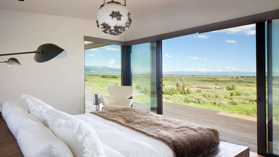 Modern bedroom with white bedding, cozy throw blanket, unique pendant light, and sliding glass doors opening to a spacious deck with panoramic countryside views.