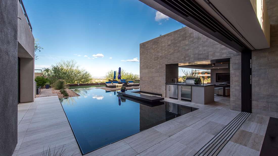 Luxury outdoor living space with infinity-edge pool, modern stone patio, built-in outdoor kitchen, and panoramic desert views under a clear blue sky.
