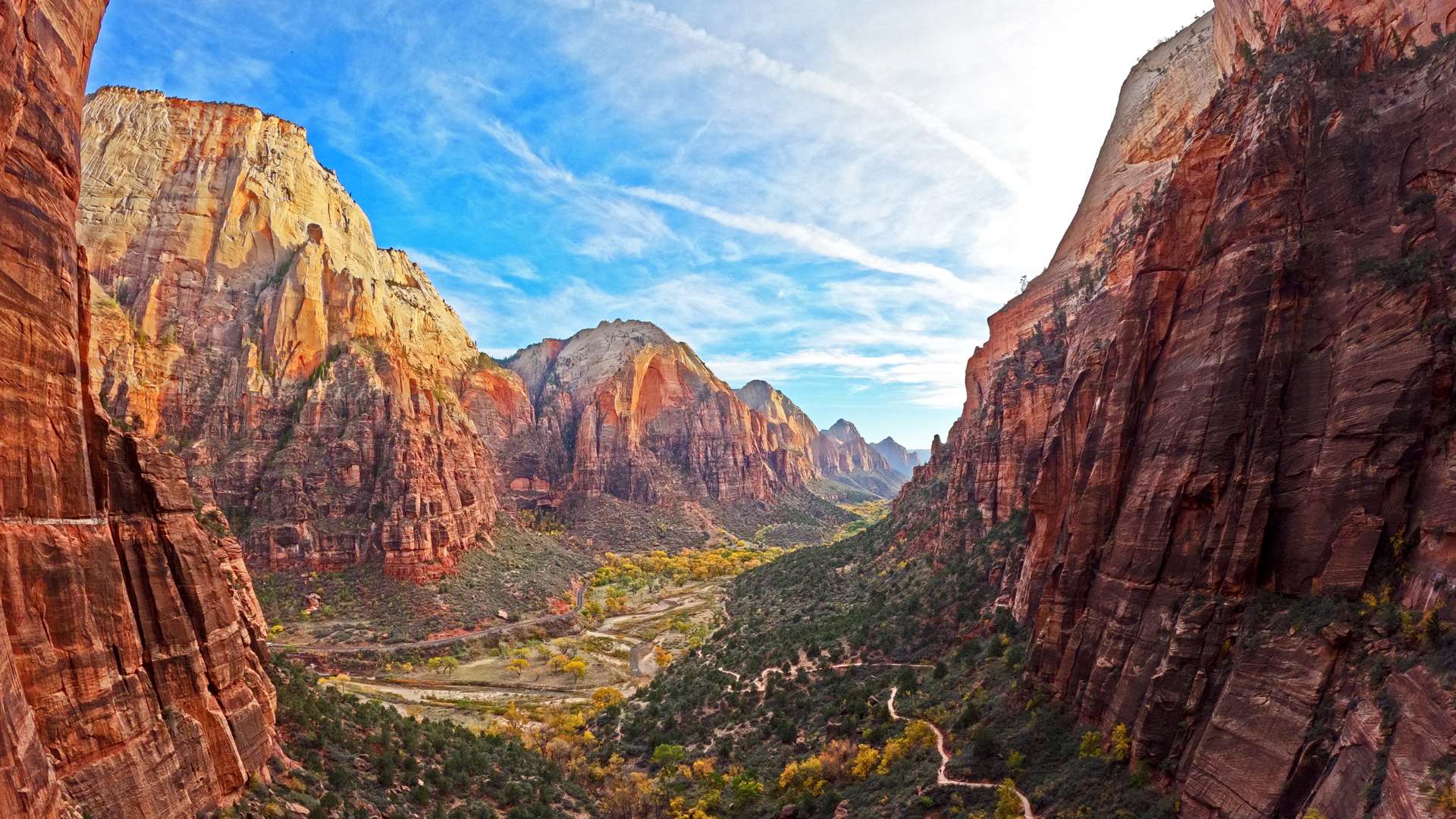   Panoramic view of a canyon with towering red cliffs, winding valley trails, and a bright blue sky.