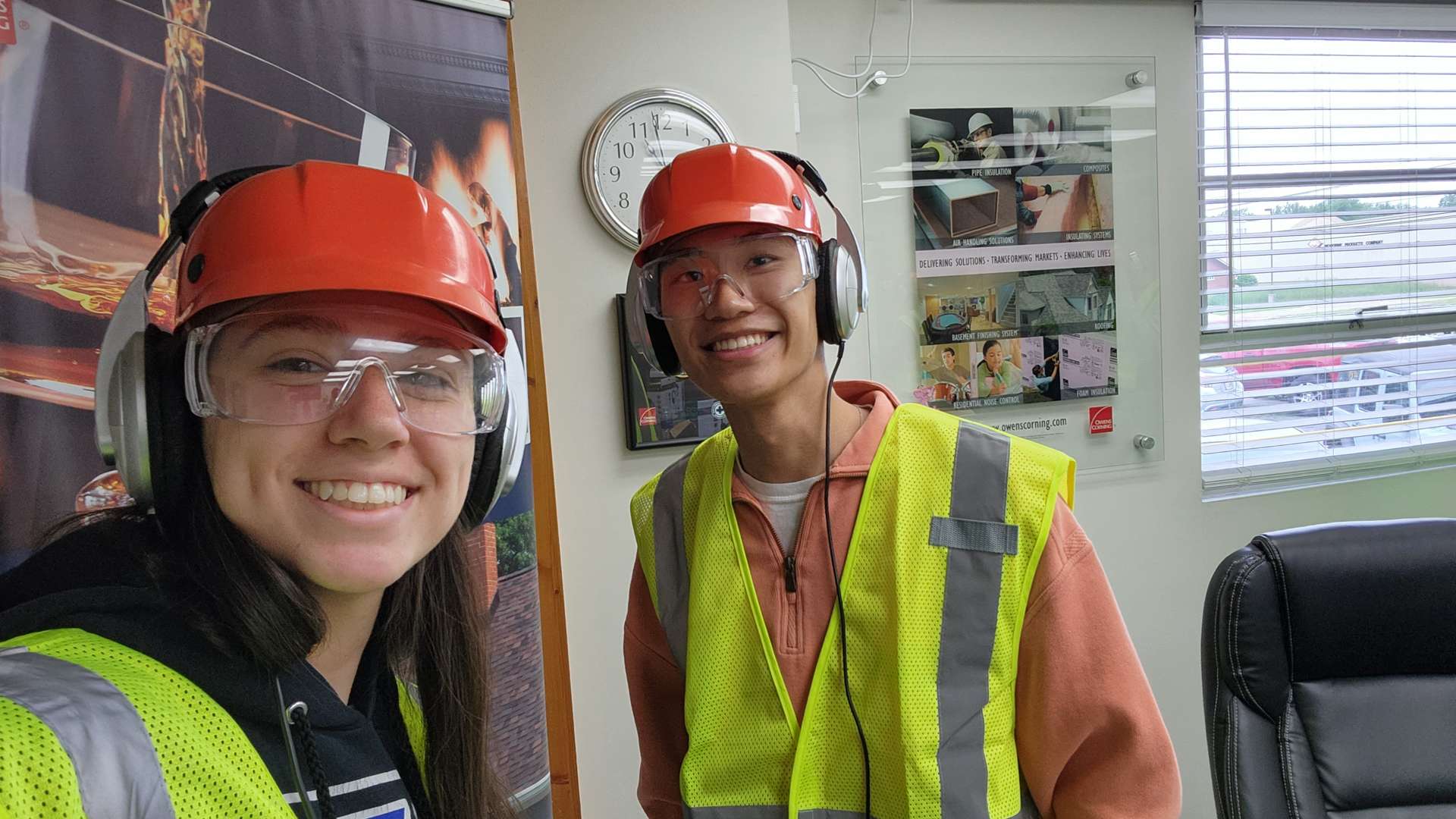 Two people wearing orange safety helmets, earmuffs, and yellow reflective vests standing inside an office with posters on the wall, a clock, and a window with blinds.