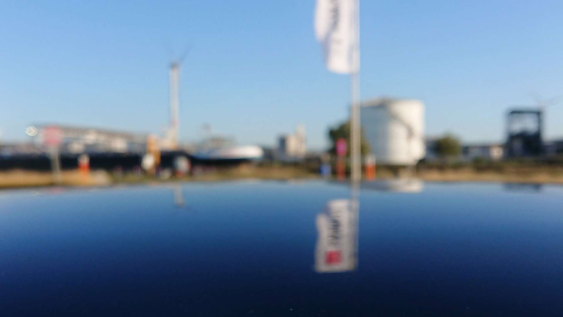 Close-up of a ladybug on a shiny black car surface with blurred industrial background, flags, and wind turbines under blue sky.