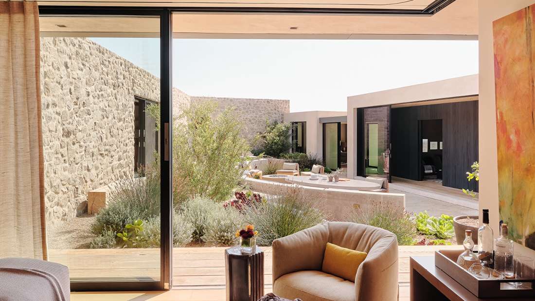 Modern indoor-outdoor living space with sliding glass doors opening to a landscaped courtyard featuring stone walls, lush greenery, and outdoor seating, viewed from a cozy lounge chair with yellow accent pillow and side table.