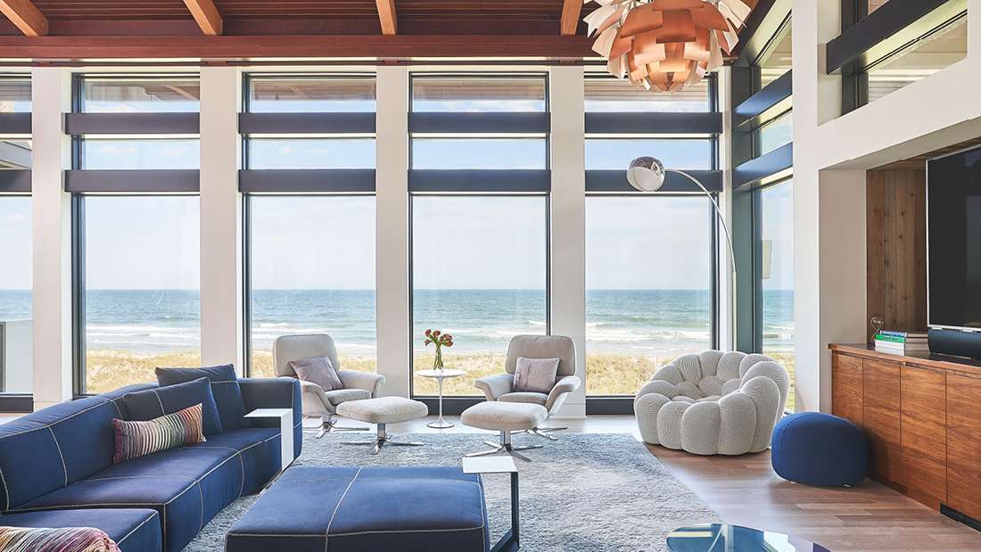 Luxury coastal living room with floor-to-ceiling windows offering panoramic ocean views, navy blue sectional sofa, modern lounge chairs, textured gray area rug, and sculptural wooden pendant light under exposed wood ceiling beams.