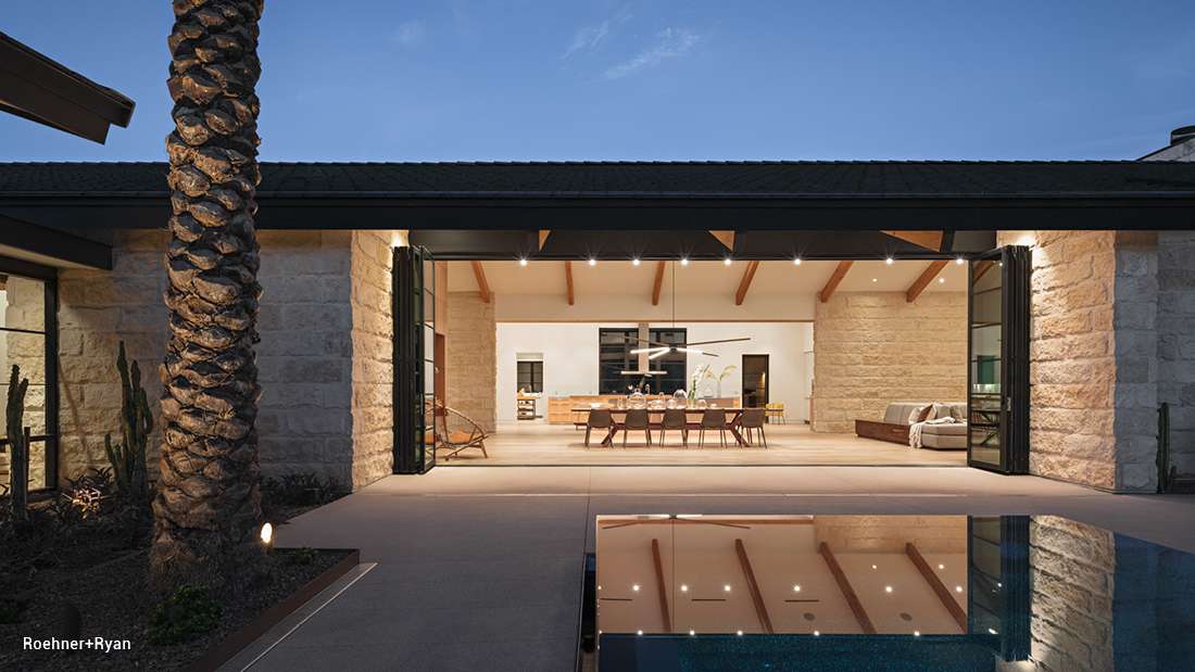 Luxury modern home exterior with folding glass doors opening to a spacious patio, illuminated dining area with vaulted ceiling and exposed wooden beams, and reflection of warm interior lights on pool surface at dusk.