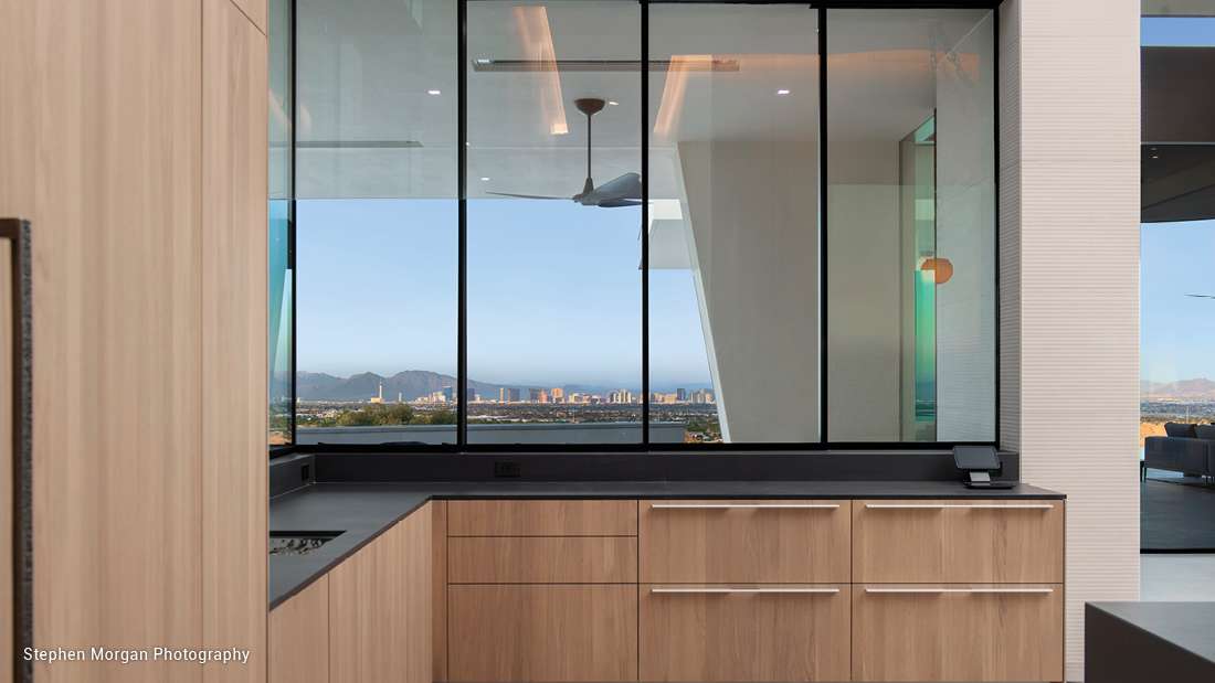 Modern kitchen with light wood cabinetry, black countertops, and large glass windows showcasing panoramic city skyline and mountain views.