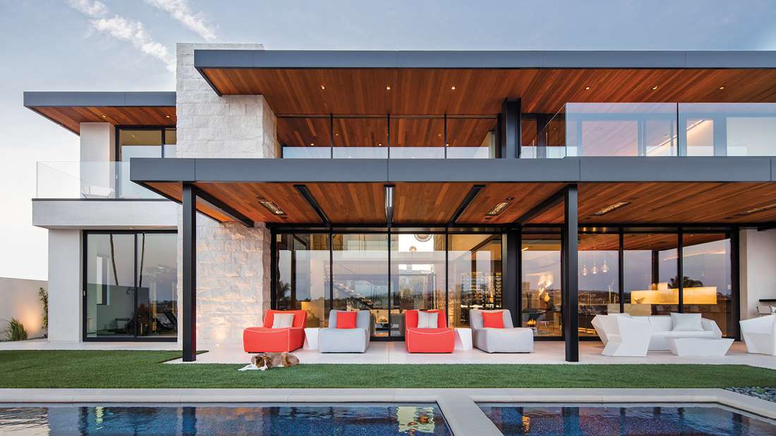 Modern two-story luxury home with floor-to-ceiling glass walls, wood-paneled ceilings, stone accent wall, outdoor lounge seating, and swimming pool in front.