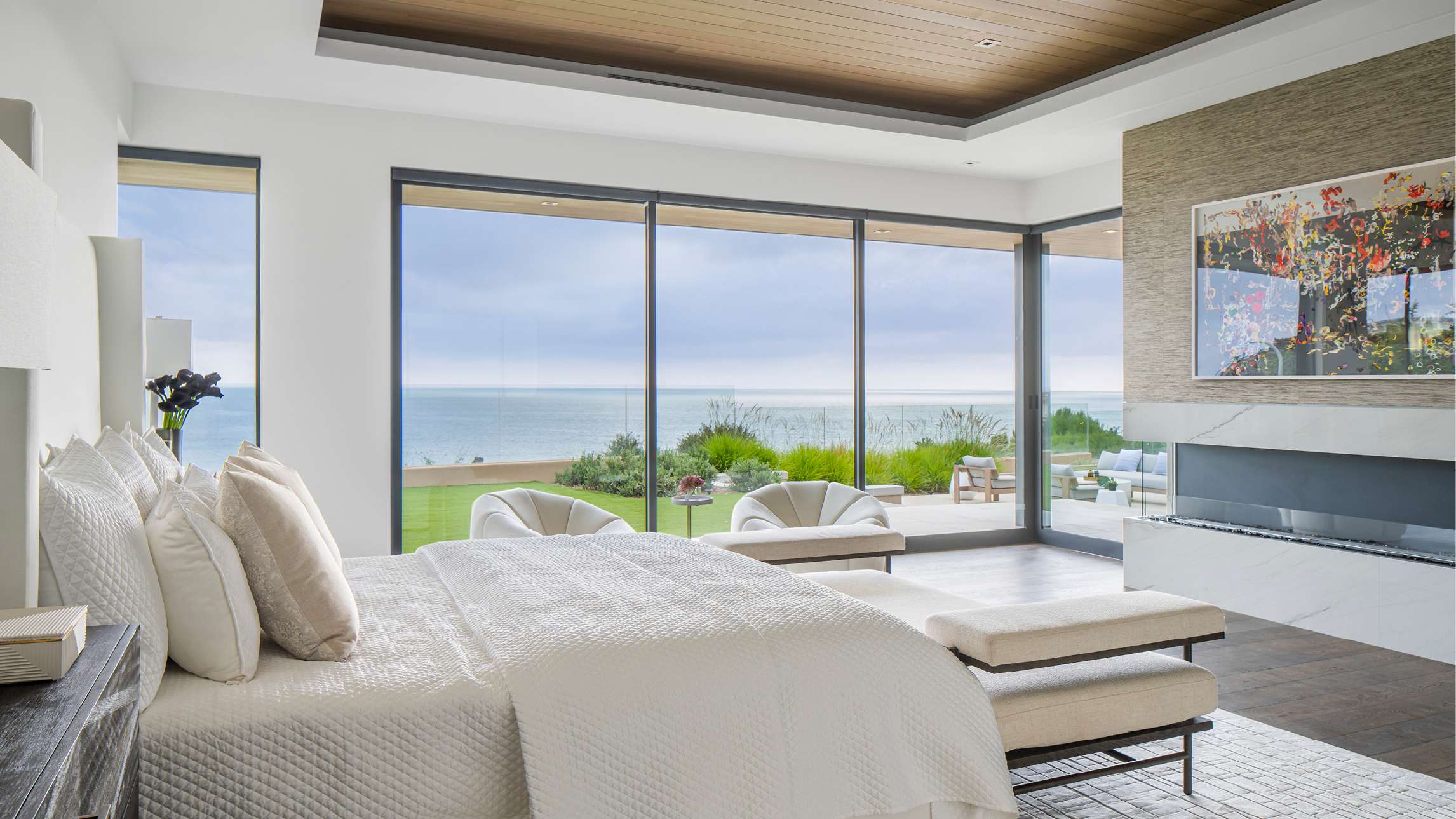 Luxury coastal bedroom with floor-to-ceiling glass walls offering panoramic ocean views, white upholstered bed with layered pillows, modern fireplace with marble surround, and wood-paneled ceiling.