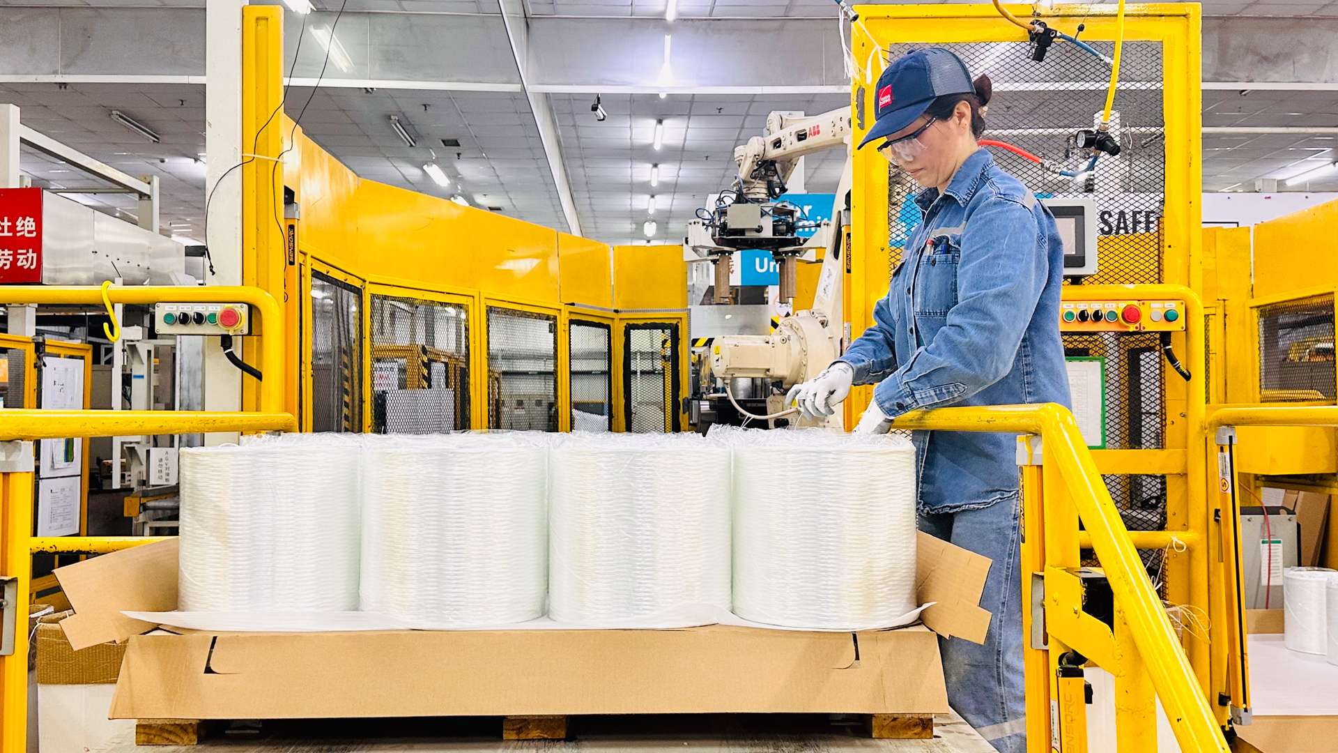 Worker in denim clothing operating machinery near large rolls of white material in a bright yellow industrial setting.