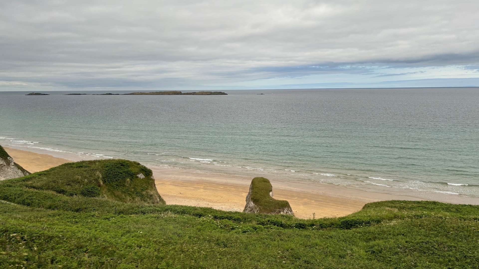 Sandy beach with gentle waves and grassy cliffs overlooking calm sea under a cloudy sky with distant islands.