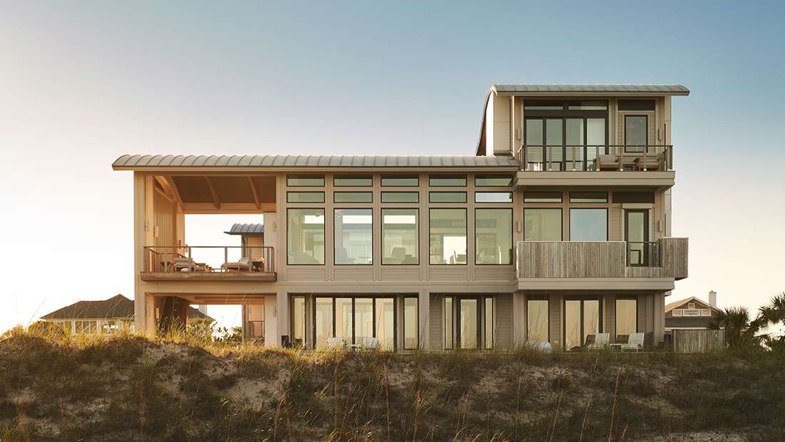 Luxury beachfront home with three stories, expansive floor-to-ceiling windows, multiple balconies with wooden railings, and open-air terraces overlooking ocean views at sunset.