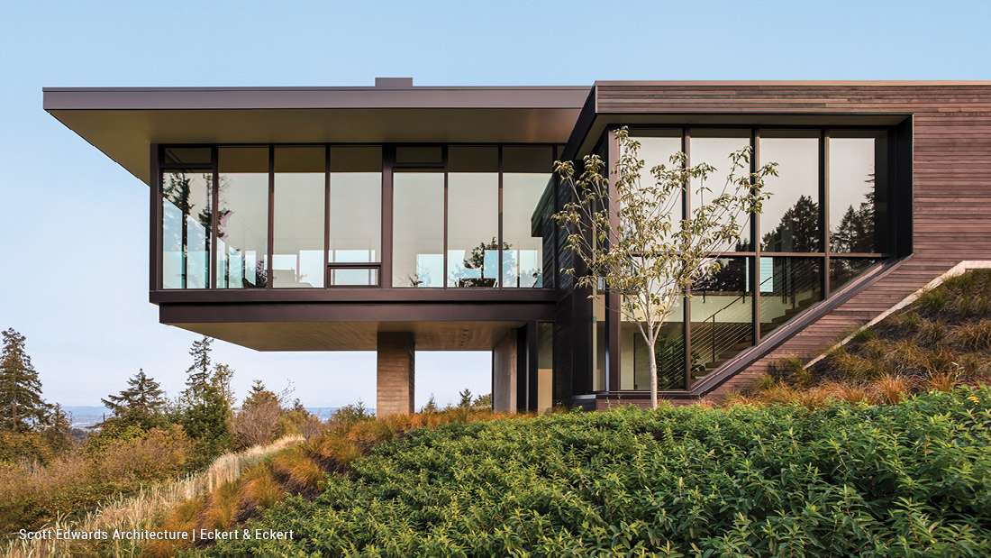 Modern hillside home with cantilevered upper level, floor-to-ceiling glass walls, flat roof design, and dark wood siding surrounded by lush greenery and panoramic valley views.