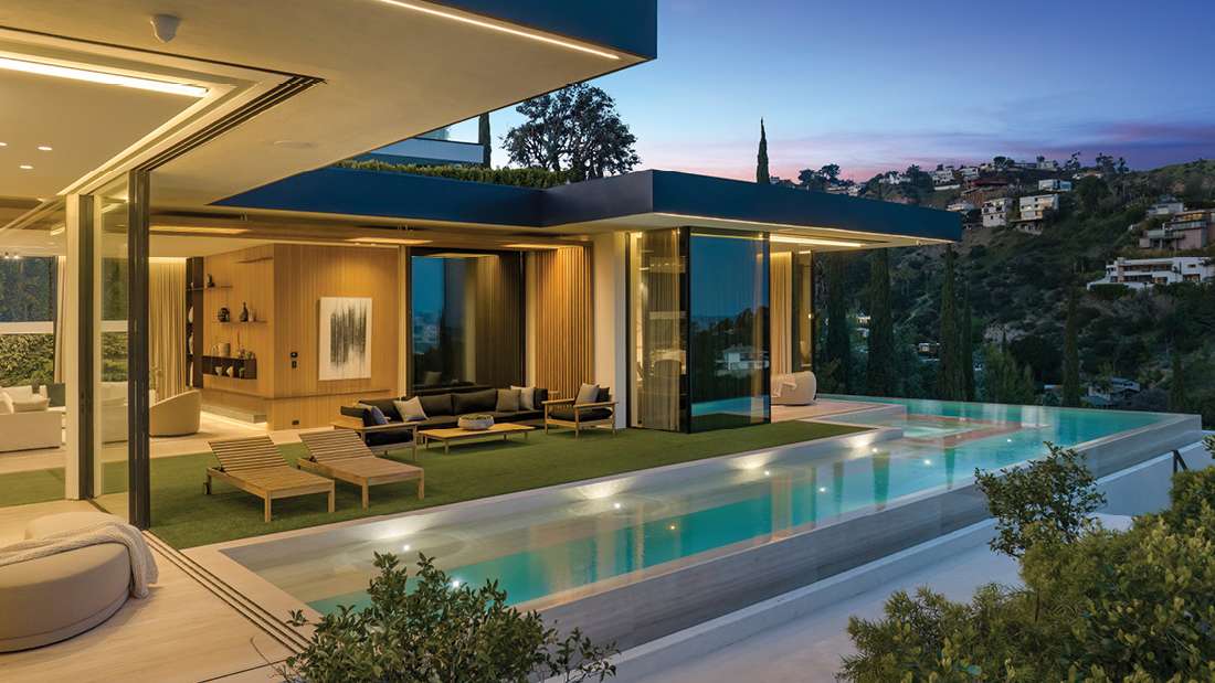 Modern luxury home with open-concept living area, floor-to-ceiling glass walls, infinity pool, outdoor lounge chairs, and hillside views at sunset