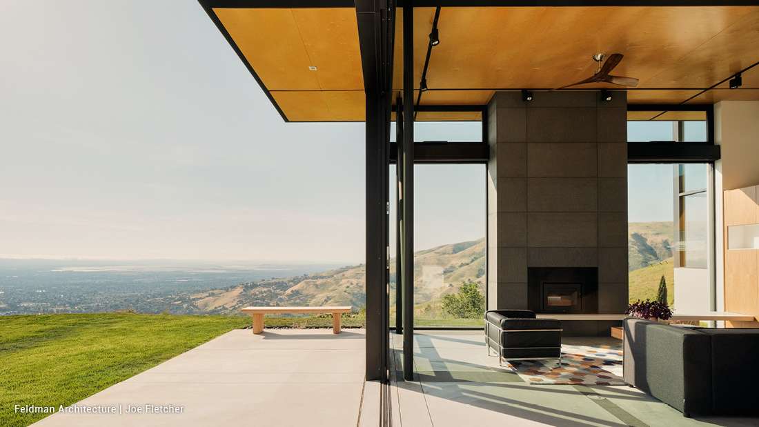 Modern hillside home with open-concept living area, large glass walls, minimalist fireplace, and panoramic valley views under a cantilevered roof.