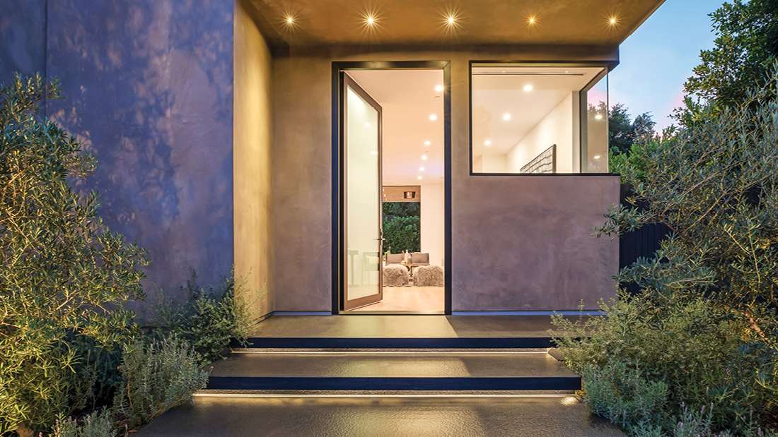 Modern home entrance with minimalist design, large pivot glass door framed in black, recessed ceiling lights, and landscaped pathway surrounded by lush greenery.