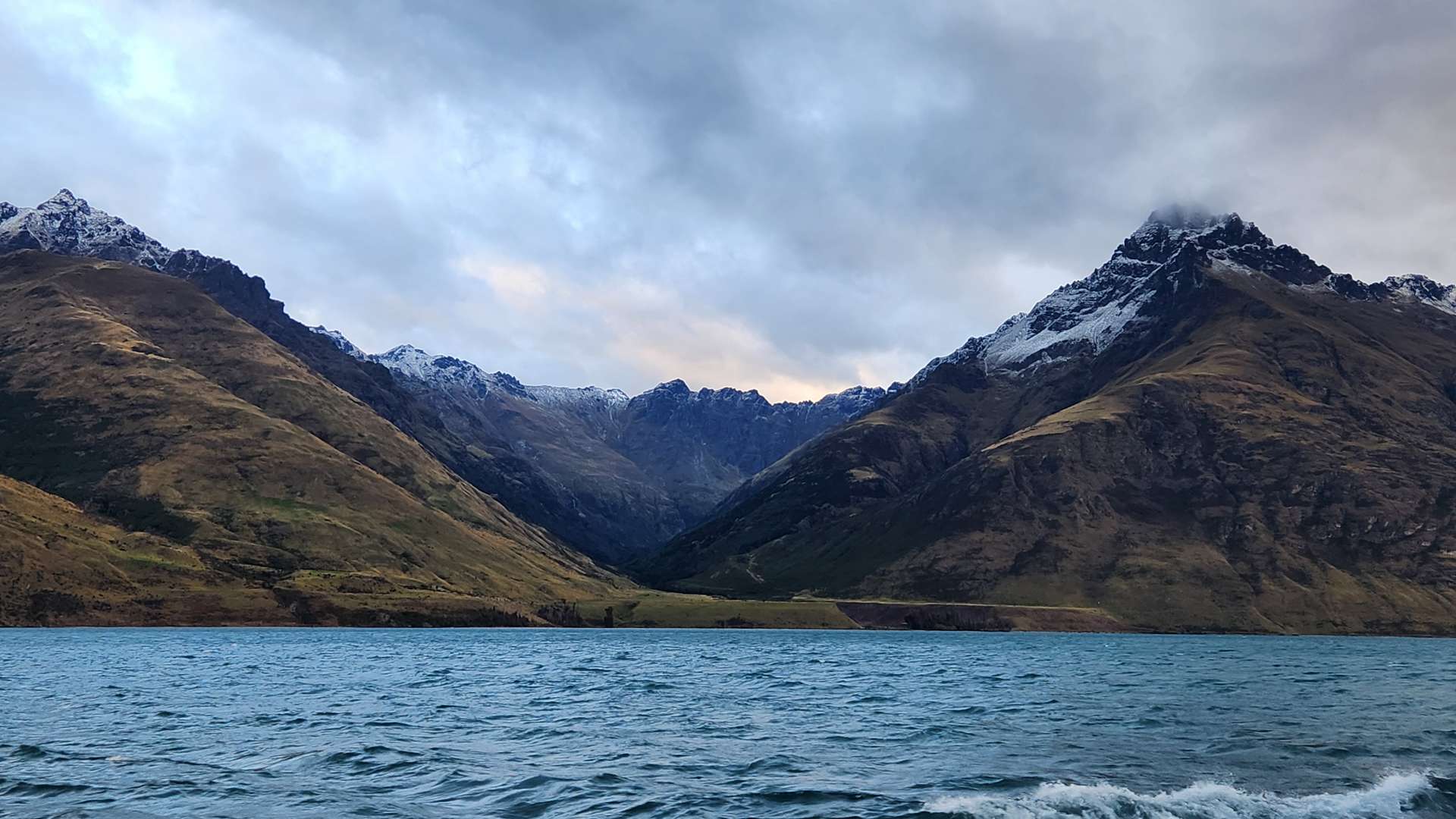   Choppy blue lake with snow-capped mountain peaks and rugged slopes under a cloudy sky at sunset.