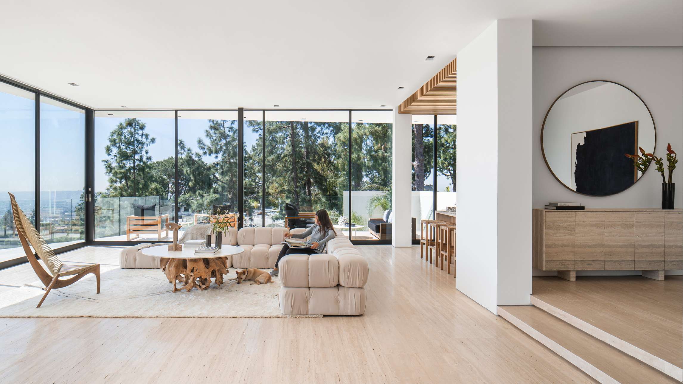 Luxury open-concept living room with floor-to-ceiling glass walls offering panoramic views of pine trees and ocean, modular beige sectional sofa, natural wood coffee table, and modern bar area with wooden stools.