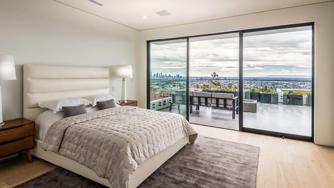 Luxury bedroom with plush upholstered bed, neutral tones, large sliding glass doors opening to a terrace with panoramic city views.