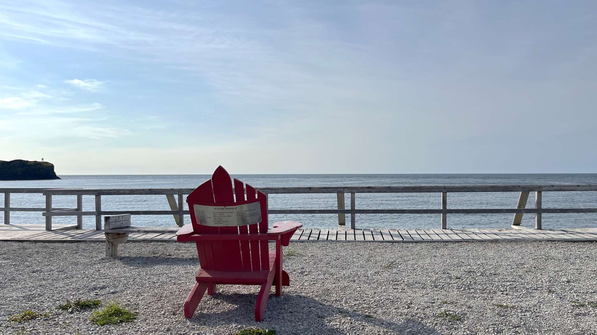 Red Adirondack chair facing a calm ocean view from a gravel beach with wooden boardwalk under a clear blue sky.