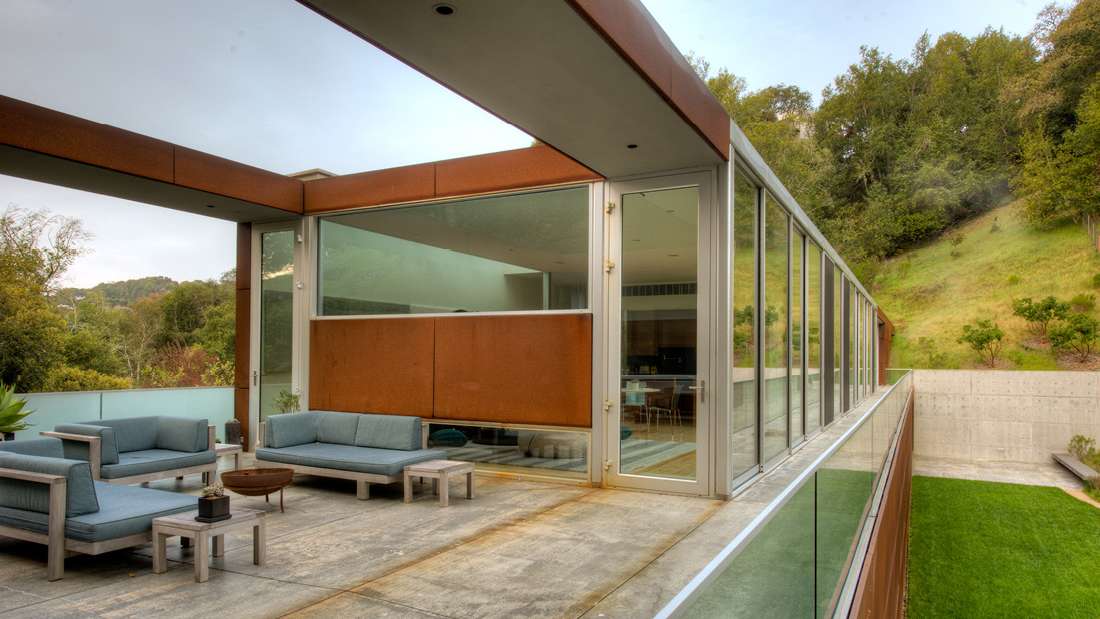 Modern outdoor terrace with concrete flooring, glass railing, and rust-colored steel frame, featuring minimalist lounge seating with blue cushions and panoramic hillside views.