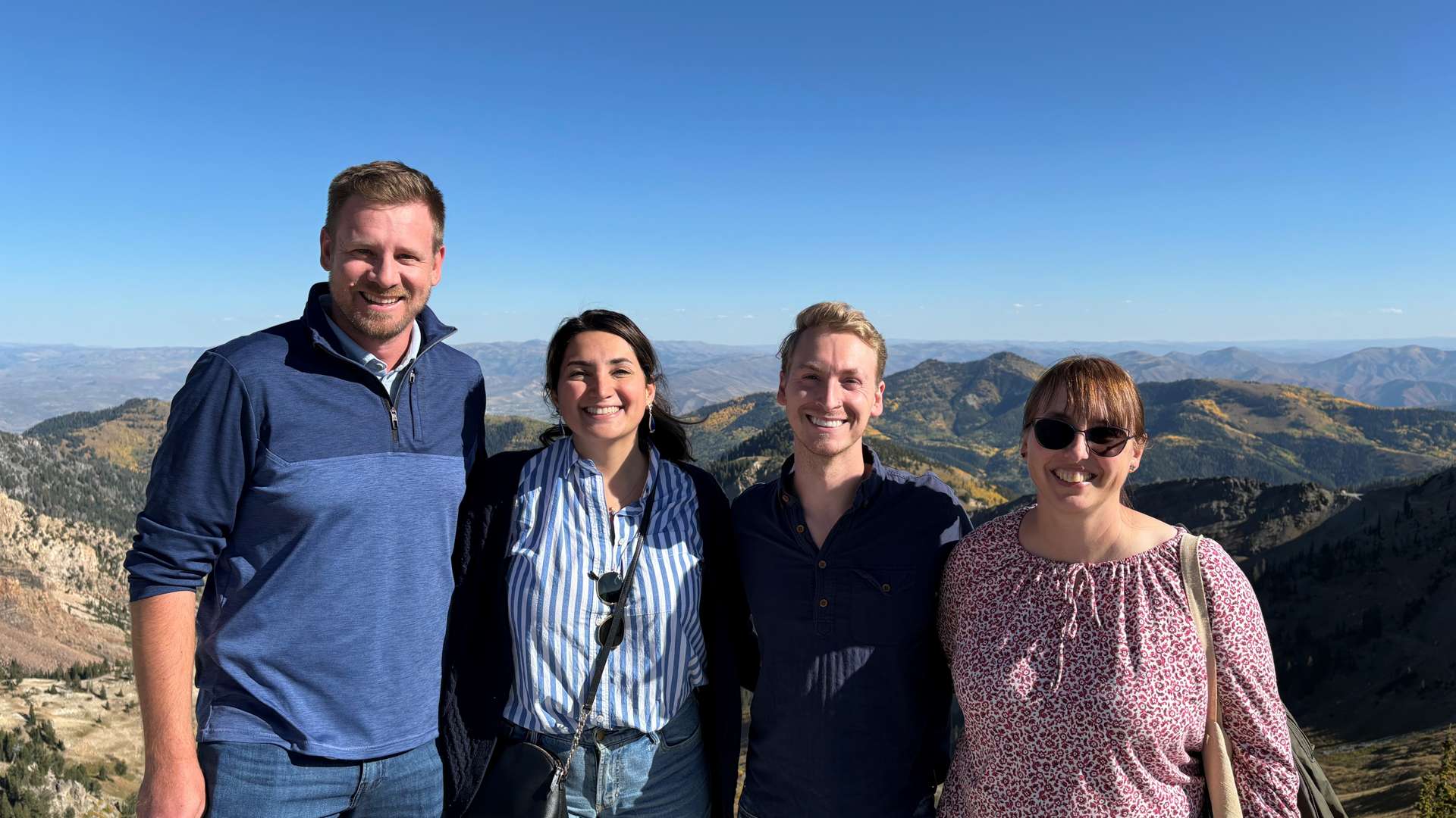 Four people standing together on a mountain overlook with rugged peaks and valleys in the background under a clear blue sky.