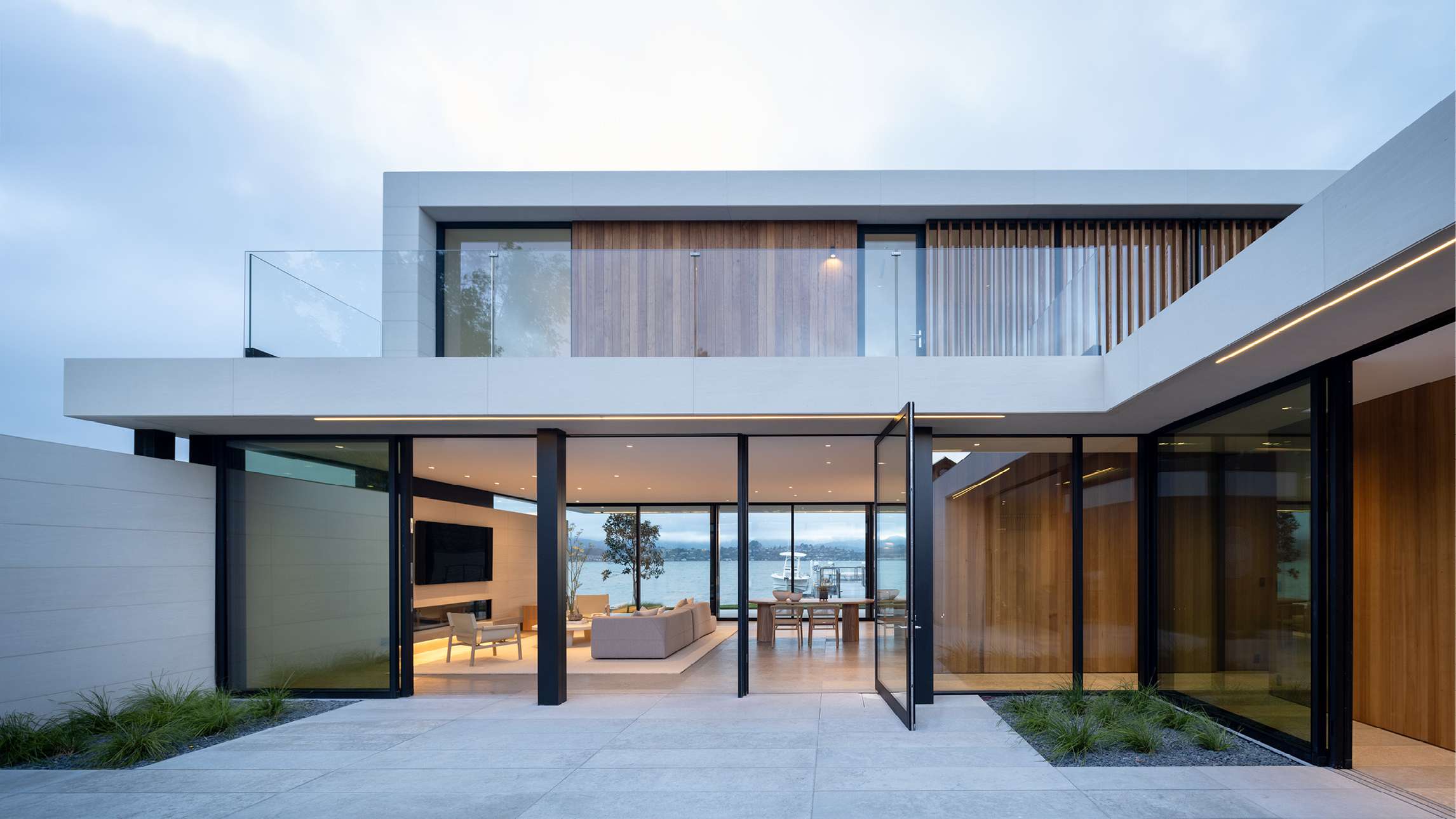 Luxury modern two-story home with minimalist white exterior, glass balcony railings, vertical wooden slats, and floor-to-ceiling glass doors opening to an outdoor terrace overlooking waterfront views.