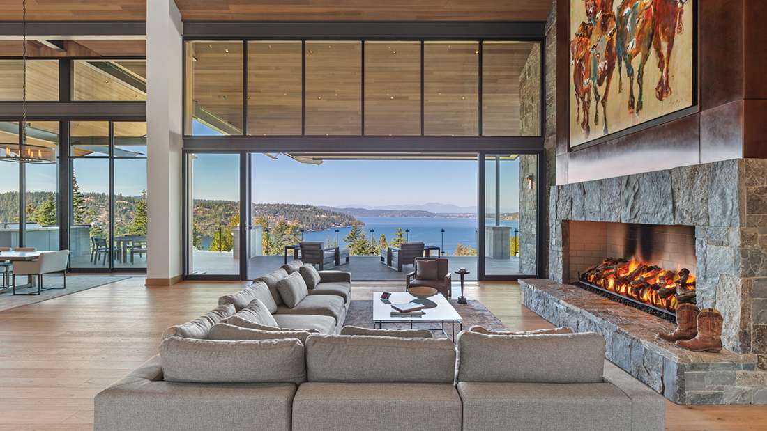 Luxury open-concept living room with floor-to-ceiling glass walls, panoramic lake views, large sectional sofa, stone fireplace with roaring fire, and modern artwork above mantel.