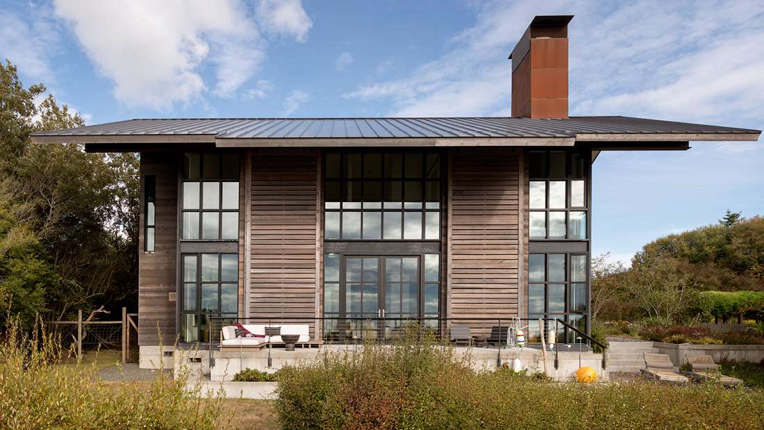 Contemporary two-story home with vertical wood siding, floor-to-ceiling glass windows, metal roof, and outdoor seating area on elevated patio, featuring a rust-colored chimney accent.